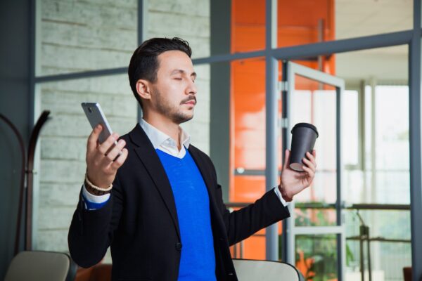 Young businessman meditating with smartphone and coffee at modern office.